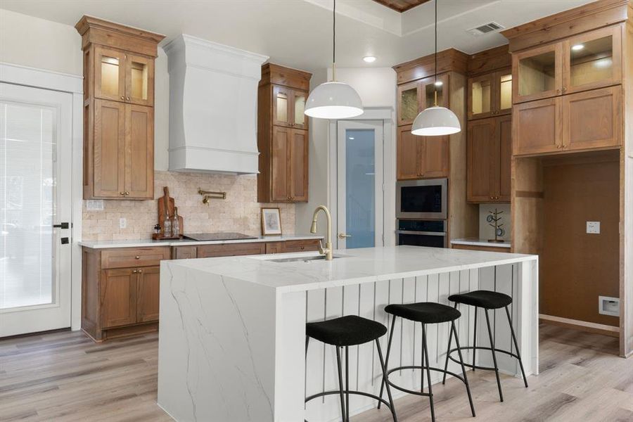 Kitchen featuring premium range hood, a kitchen island with sink, sink, built in microwave, and decorative light fixtures