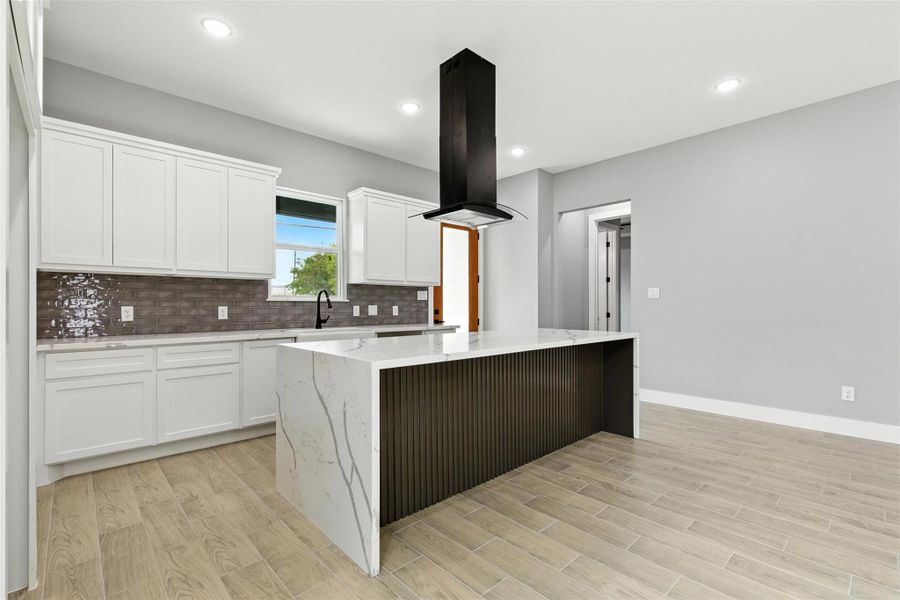 Modern kitchen with sleek white cabinetry, a large island with a marble countertop, and a stylish black range hood. The space features light flooring, grey walls, and ample natural light.