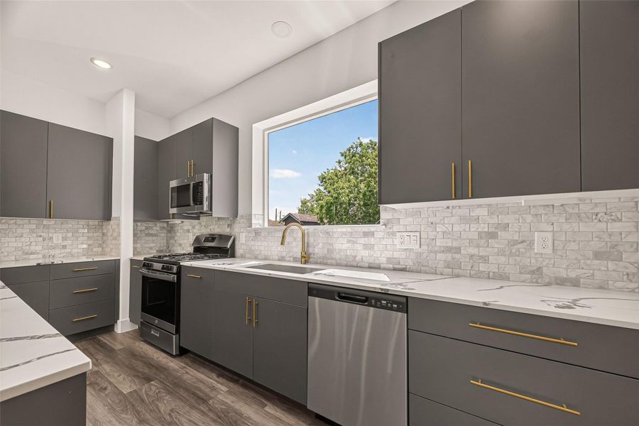 This modern kitchen features sleek gray cabinets with gold handles, stainless steel appliances, and a large window that brings in natural light. The marble backsplash and countertops add a touch of elegance, while the vinyl flooring provides warmth. This modern kitchen features sleek gray cabinets with gold handles, stainless steel appliances, and a large window that brings in natural light. The marble backsplash and countertops add a touch of elegance, while the vinyl flooring provides warmth.
