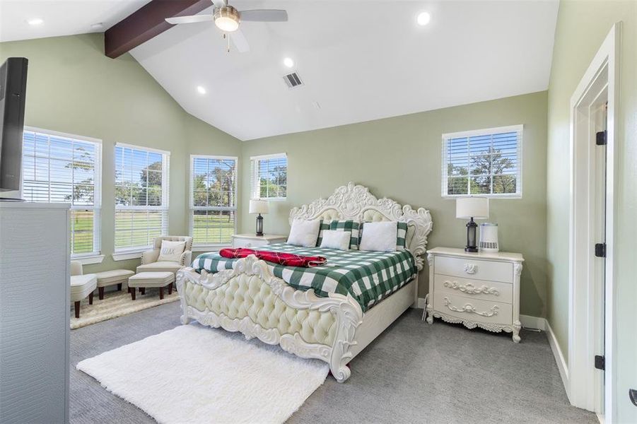 Bright and spacious bedroom with vaulted ceilings and large windows offering plenty of natural light. Features a decorative bed, matching nightstands, and a cozy seating area. Neutral tones create a relaxing ambience.