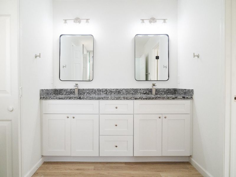 This photo shows a modern bathroom with dual sinks, white cabinetry, granite countertops, and sleek fixtures. The space is well-lit with two mirrors and contemporary light fixtures, featuring a neutral color palette and wood-look flooring.