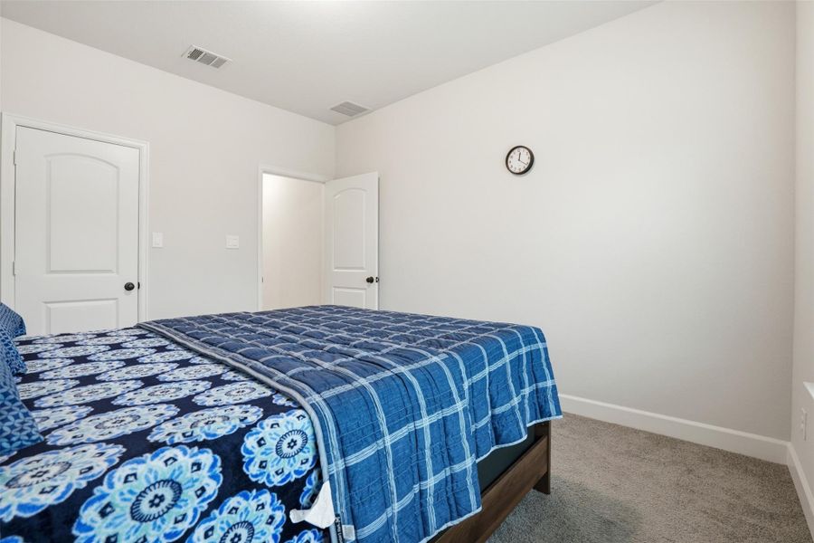 This is a cozy bedroom featuring a simple design. It has light-colored walls, a carpeted floor, and a bed with blue patterned bedding. A small wall clock is mounted above, and there are two white doors leading to adjacent areas. This is a cozy bedroom featuring a simple design. It has light-colored walls, a carpeted floor, and a bed with blue patterned bedding. A small wall clock is mounted above, and there are two white doors leading to adjacent areas.