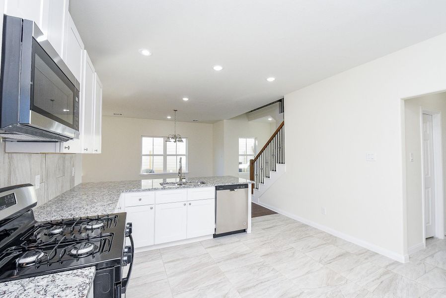 Modern kitchen with white cabinets, granite countertops, stainless steel appliances, and an adjoining open living space with stairs. Clean, bright interior.
