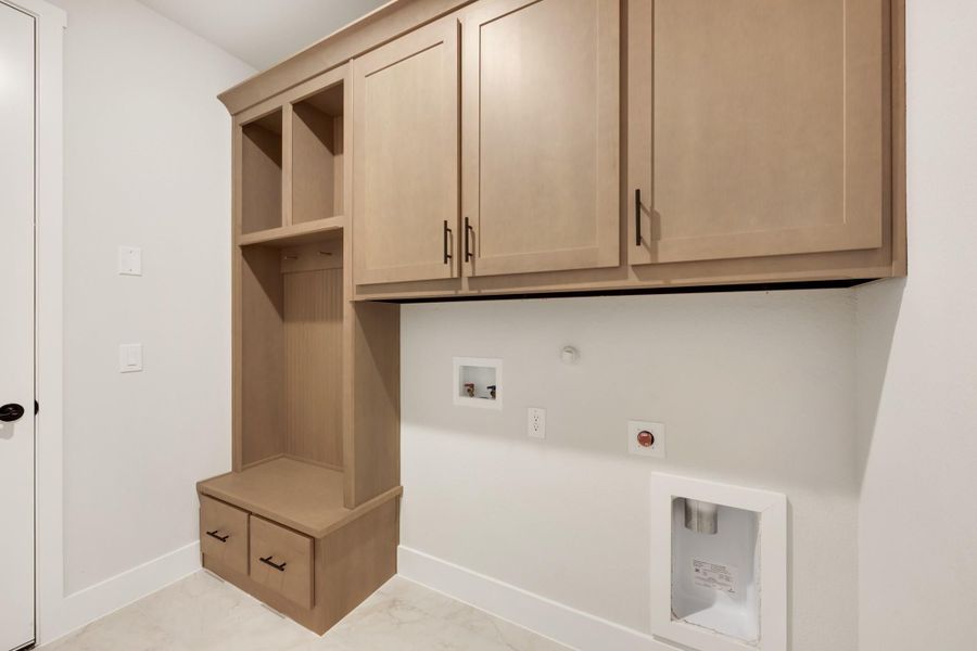 Laundry or mudroom with light wood upper cabinets, open cubbies, bench with drawers, and washer hookups on white walls.