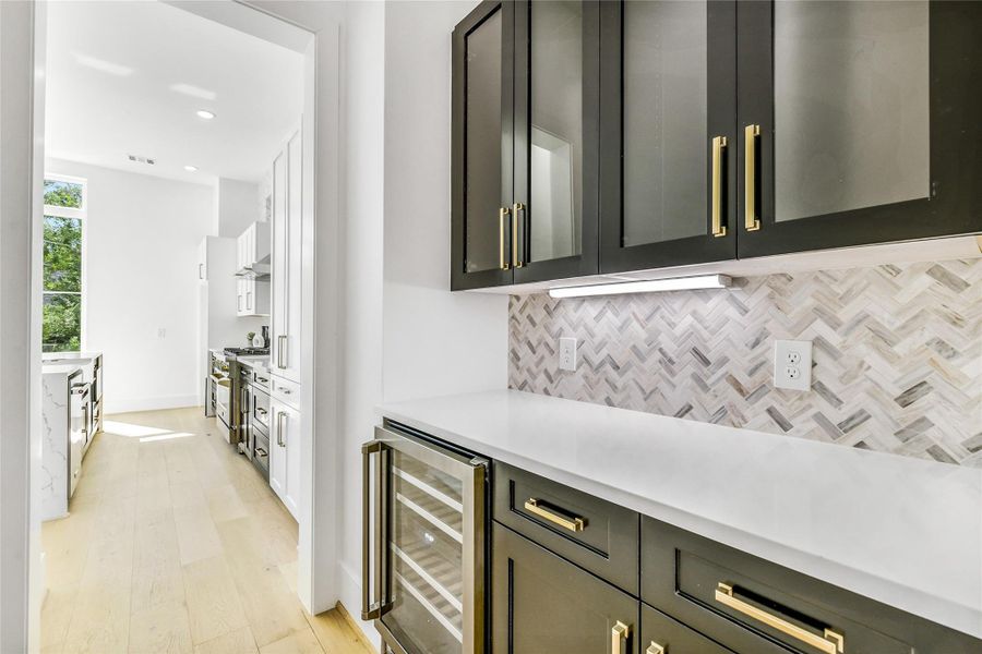 The butler’s pantry offers a stunning transition between dining and kitchen, featuring custom cabinetry in a contrasting midnight finish with soft-close drawers, brass pulls, quartz countertops, and a herringbone marble backsplash. Integrated under-cabinet lighting and a built-in wine fridge complete this stylish yet highly functional prep space—ideal for entertaining or late-night studybreaks.