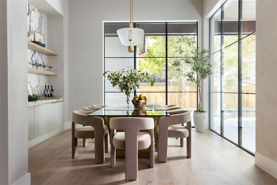 Expansive RAM windows in the dining room overlook the covered patio and pool, while a stylish dry bar featuring open shelving and reeded cabinet doors completes this serene, oasis-like setting. Expansive RAM windows in the dining room overlook the covered patio and pool, while a stylish dry bar featuring open shelving and reeded cabinet doors completes this serene, oasis-like setting.