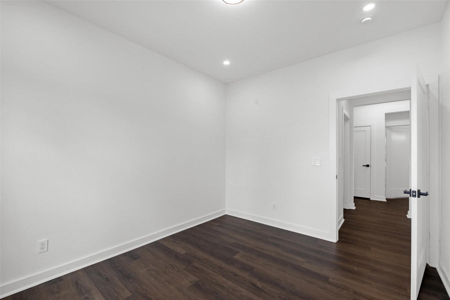 Brilliant bedroom with white walls and dark hardwood flooring. The space features recessed lighting and an open door leading to a hallway, creating a clean and inviting atmosphere.