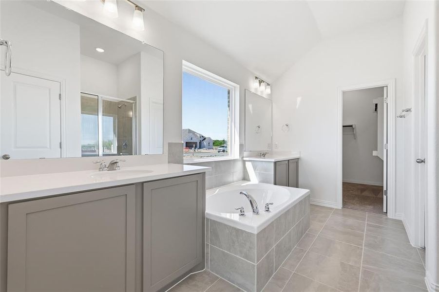 Spacious bathroom featuring a built-in tub with tiled surround, dual vanities with white countertops and undermount sinks, a large window, a separate shower with glass enclosure, and vaulted ceilings