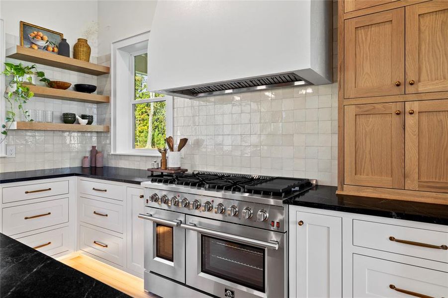 Kitchen with custom range hood, range with two ovens, open shelves, decorative backsplash, and dark stone countertops