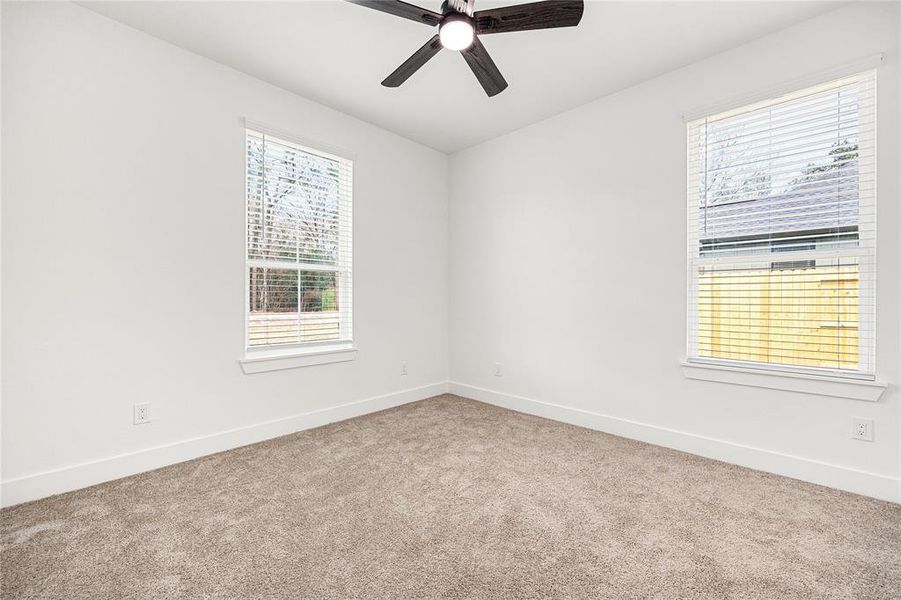 A bright, cozy room with neutral carpet and fresh white walls. Features two large windows providing natural light and a modern ceiling fan for comfort.