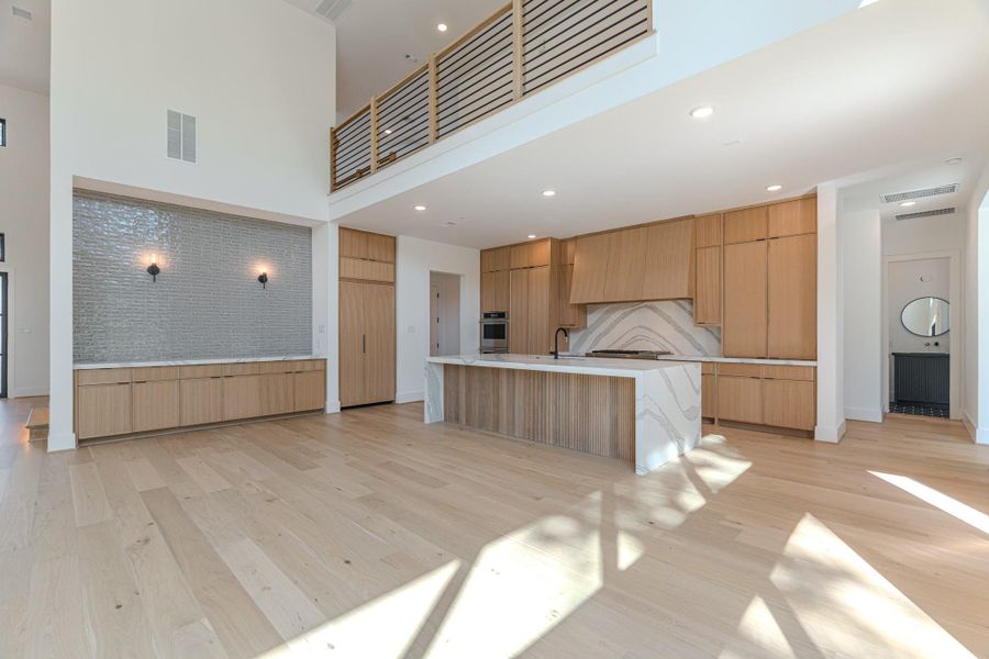 This spacious, modern kitchen features light wood cabinetry, a large island with a marble-patterned countertop, and high ceilings. The open layout includes a textured accent wall with built-in lighting, offering a stylish and functional space. Natural light fills the room, enhancing the airy feel.