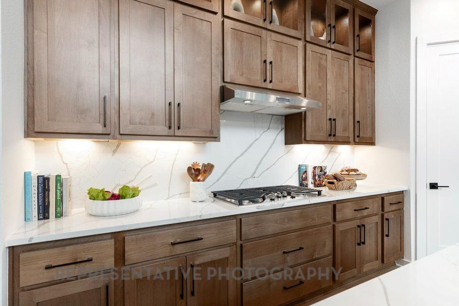 Kitchen featuring tasteful backsplash, glass insert cabinets, under cabinet range hood, light stone counters, and stainless steel gas cooktop Kitchen featuring tasteful backsplash, glass insert cabinets, under cabinet range hood, light stone counters, and stainless steel gas cooktop