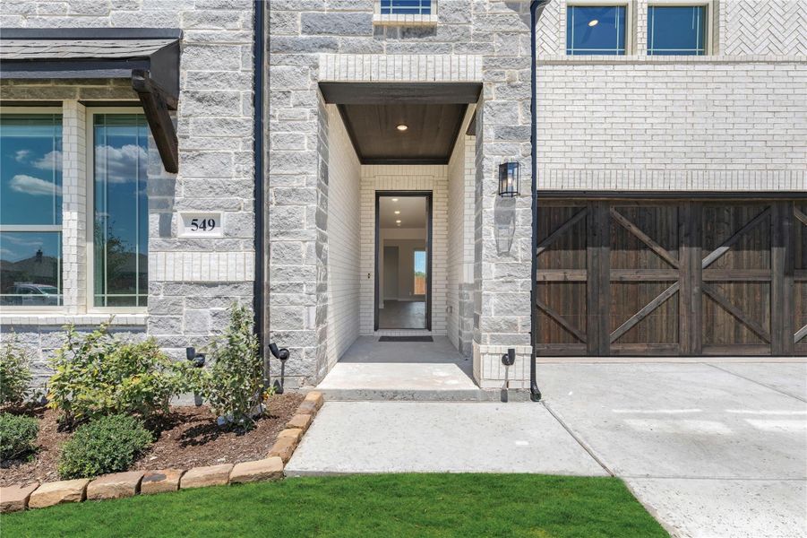 Front entry of stone and brick home with covered porch, dark wood garage door, and landscaped yard
