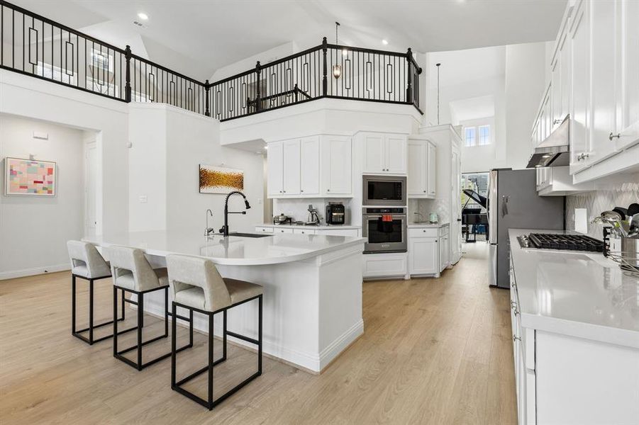 Spacious kitchen featuring an extended island with a black faucet, light wood-finish flooring, white cabinetry, stainless steel appliances, and a vaulted ceiling with an open black metal railing