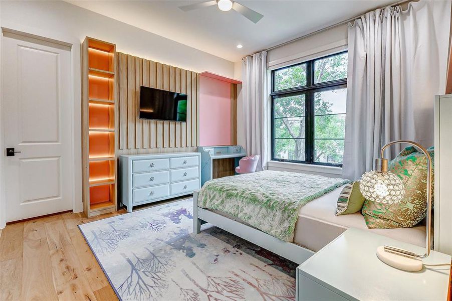 This fifth cozy bedroom features a modern design with wooden flooring, a large window for natural light, and elegant curtains. It includes a stylish bed with a matching nightstand, a built-in desk, and a wall-mounted TV, complemented by warm custom LED lighting and decorative accents.