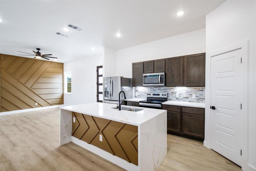 Kitchen featuring stainless steel appliances, a kitchen island with sink, dark wood finish cabinets, light wood finished floors, and light stone countertops