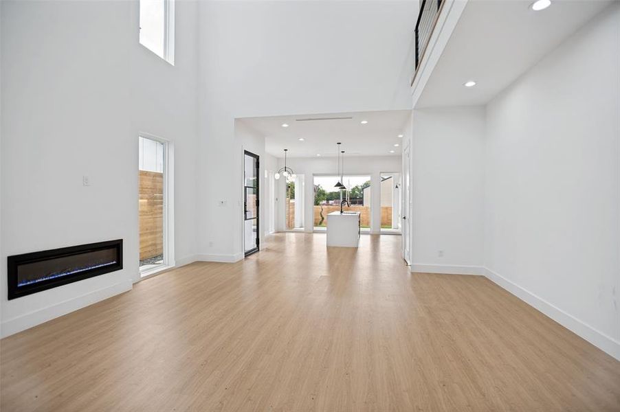 Open-concept living space featuring high ceilings, wood-finish flooring, a linear electric fireplace, recessed lighting, and a modern kitchen island with pendant lighting