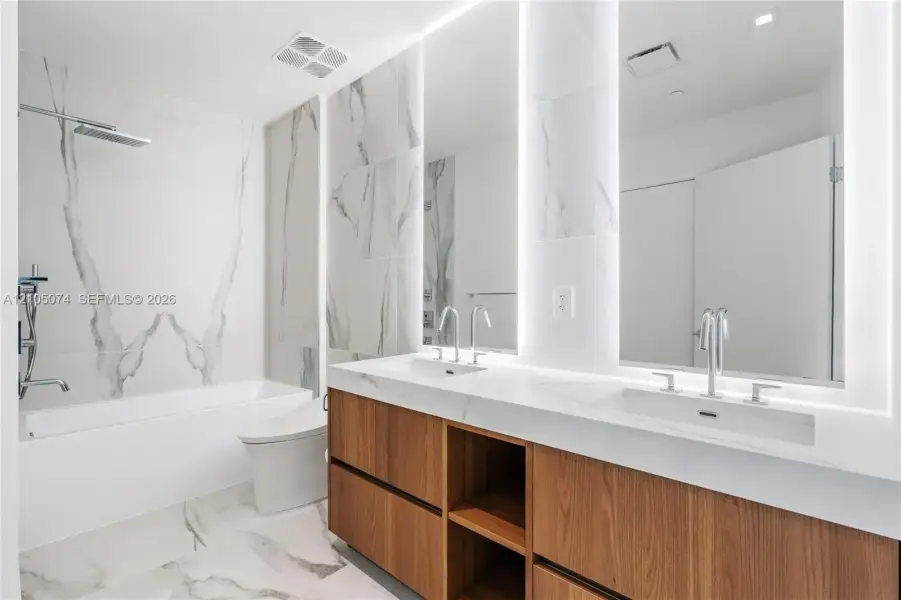 Dual-sink vanity with wood-finish cabinetry, quartz countertops, large-format tiled wall surround, undermount tub, and integrated LED mirror lighting