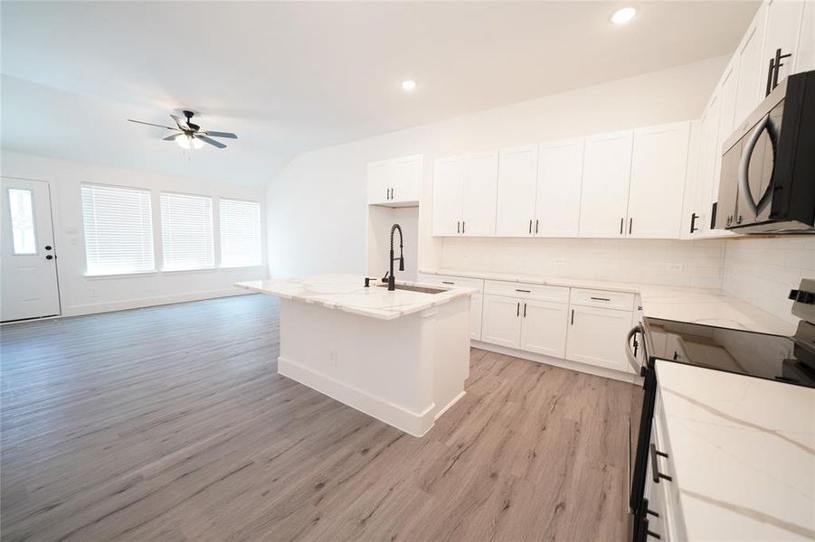 Kitchen featuring appliances with stainless steel finishes, decorative backsplash, a kitchen island with sink, light wood-style floors, and light stone countertops