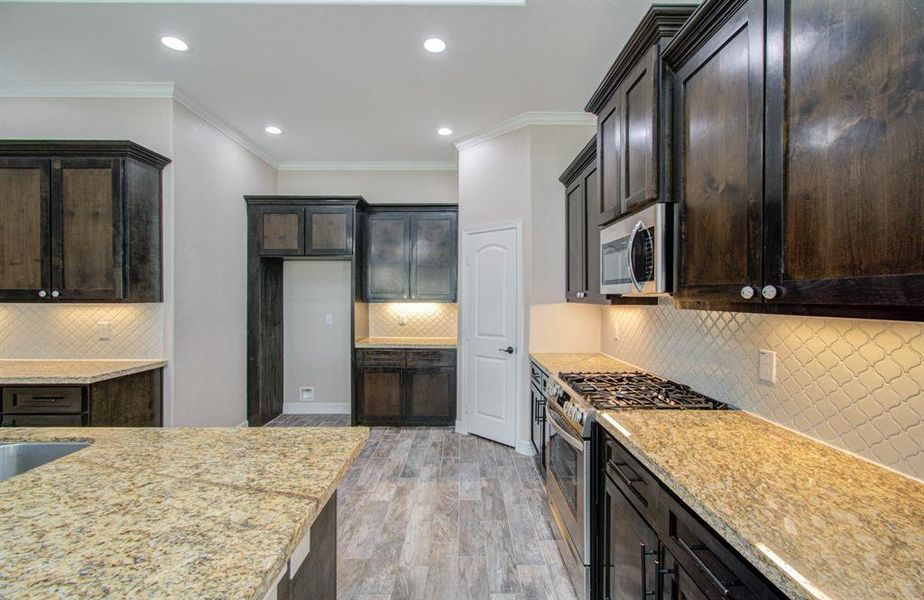 This modern kitchen features rich dark wood cabinetry, granite countertops, stainless steel appliances, and stylish backsplash tiles. The room is well-lit with recessed ceiling lights and offers ample storage and counter space.