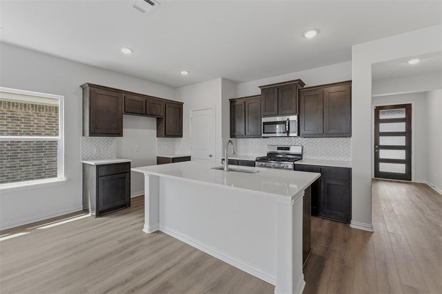 Contemporary kitchen featuring a large central island with a sink, dark wood-finish cabinetry, stainless steel appliances, a white herringbone tile backsplash, and wood-finish flooring
