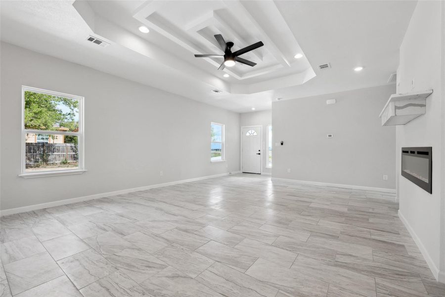 Unfurnished living room with a raised ceiling, plenty of natural light, recessed lighting, a ceiling fan, and a glass covered fireplace Unfurnished living room with a raised ceiling, plenty of natural light, recessed lighting, a ceiling fan, and a glass covered fireplace