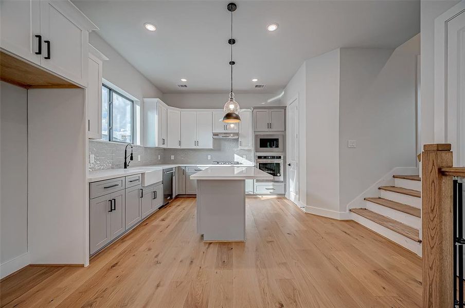 This modern kitchen features sleek white and gray cabinetry, a central island with pendant lighting, and stainless steel appliances. The space is bright and airy with natural wood flooring and a large window for natural light. A staircase is adjacent, adding a stylish touch to the open layout.