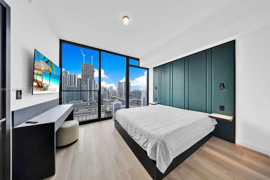 Floor-to-ceiling windows with urban skyline backdrop, custom floor-to-ceiling upholstered teal headboard, built-in desk with integrated shelving, and wide-plank wood-finish flooring Floor-to-ceiling windows with urban skyline backdrop, custom floor-to-ceiling upholstered teal headboard, built-in desk with integrated shelving, and wide-plank wood-finish flooring