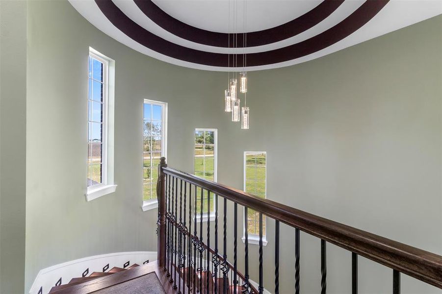 This photo showcases a curved staircase with elegant wrought iron railings, leading up to a landing with large, vertical windows that allow ample natural light. The ceiling features modern pendant lighting and decorative wood accents, creating a sophisticated ambiance.