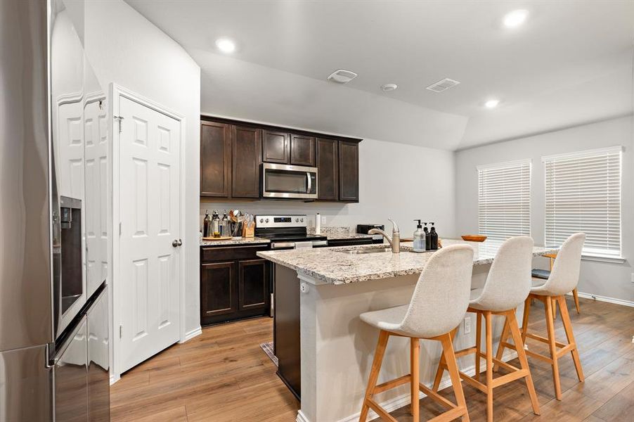 Kitchen with appliances with stainless steel finishes, light stone countertops, dark brown cabinetry, a breakfast bar, and a kitchen island with sink Kitchen with appliances with stainless steel finishes, light stone countertops, dark brown cabinetry, a breakfast bar, and a kitchen island with sink