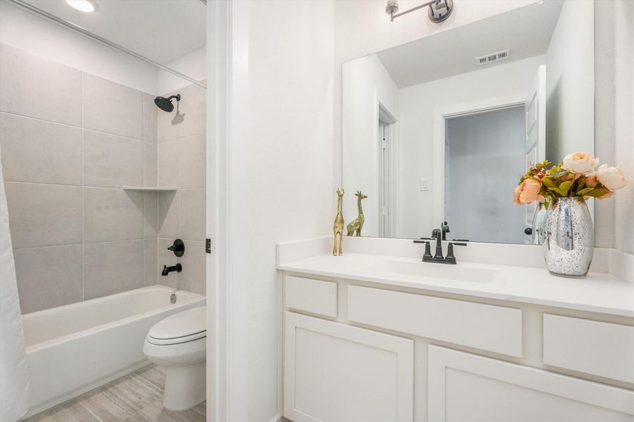 This bathroom features a modern design with a large mirror above a white vanity. It includes a bathtub with a sleek, tiled shower area and black fixtures. The space is bright and inviting, accented with decorative elements like a vase of flowers.
