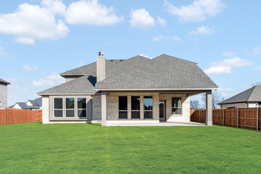 Rear exterior of brick home with covered patio, large windows, chimney, and fenced backyard with green lawn