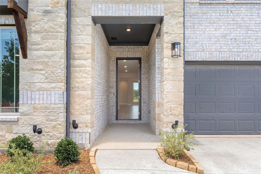 Home entrance with stone exterior, glass front door, covered entry, and gray garage door nearby Home entrance with stone exterior, glass front door, covered entry, and gray garage door nearby