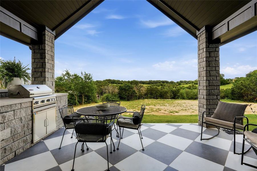 Covered patio featuring a checkerboard-patterned floor, stone columns, and an outdoor kitchen with a built-in grill