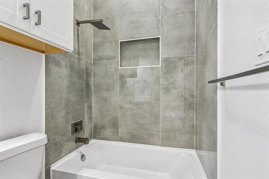 Bathtub with a rainfall shower head, stone-look tile surround, and built-in shower niche