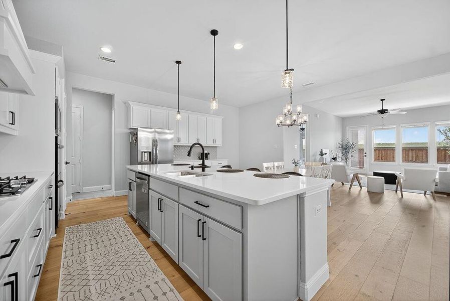 Open-concept kitchen featuring an expansive island with a built-in sink, stainless steel appliances, white cabinetry with matte black hardware, wood-finish flooring, and pendant lighting