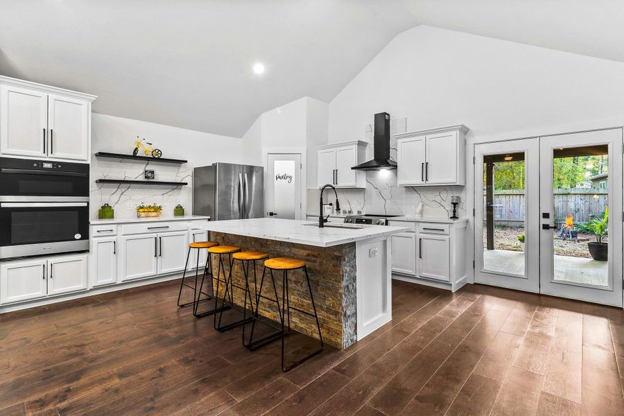 This modern kitchen features high ceilings, sleek white cabinetry, and stainless steel appliances. It boasts a large center island with seating and a stone accent, wood flooring, and French doors leading to a backyard, providing a bright and inviting space.