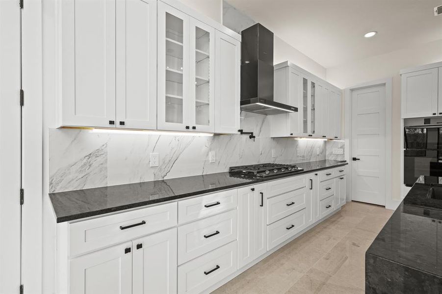 Kitchen with stainless steel gas stovetop, white cabinetry, dark stone countertops, and wall chimney exhaust hood