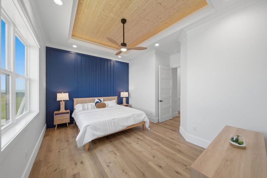 This bright bedroom features a stylish wooden ceiling with a fan, a bold blue accent wall, and ample natural light from large windows. The space includes a modern bed with matching side tables on a hardwood floor.