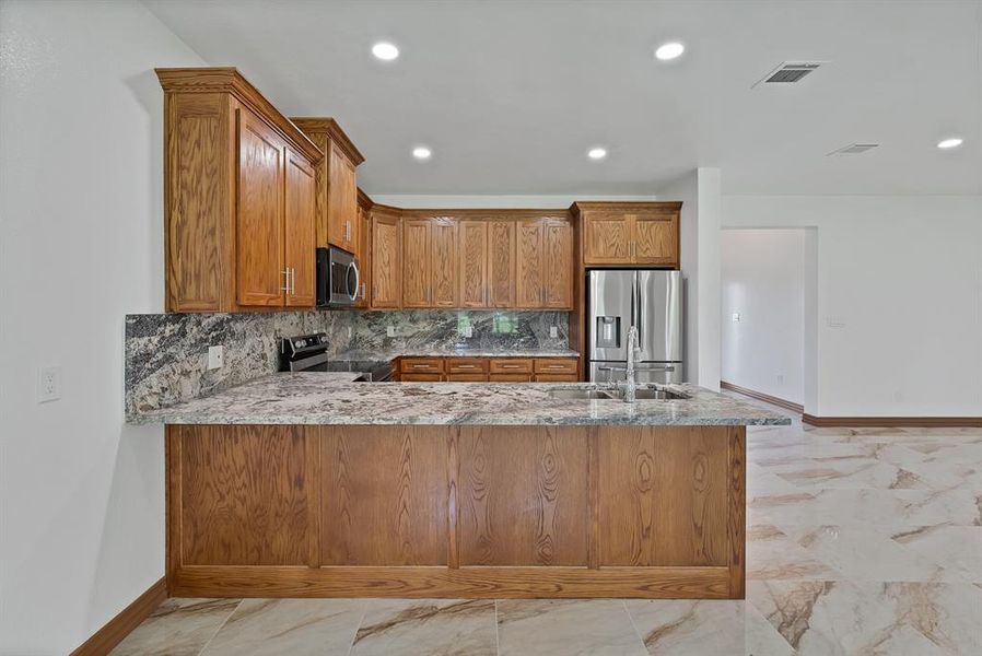 Kitchen with brown cabinetry, light stone counters, stainless steel appliances, a peninsula, and recessed lighting