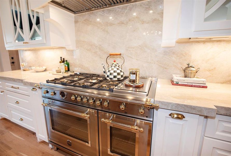 This kitchen features a high-end stainless steel stove with multiple burners, set against a sleek marble backsplash. The countertops are elegant and spacious, complemented by stylish white cabinetry with brass handles, creating a luxurious and functional cooking space.