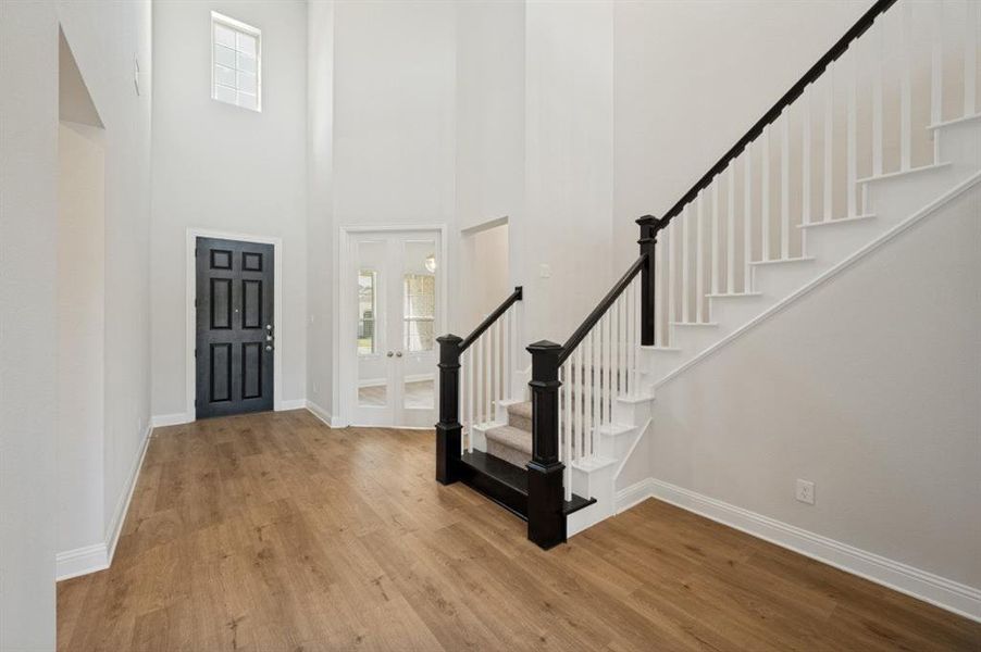 Two-story entryway featuring wood-finish flooring, a dark paneled front door, white double doors with glass panels, a staircase with dark newel posts and handrails, and a high window