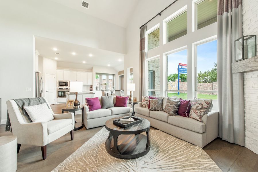 Open-concept living room with two-story windows, gray sofas, and hardwood floors open to kitchen Open-concept living room with two-story windows, gray sofas, and hardwood floors open to kitchen