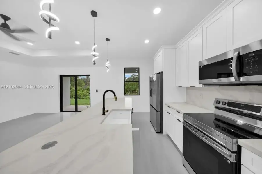 Kitchen featuring white shaker-style cabinetry, a stone-finish island with a built-in farmhouse sink, stainless steel appliances, matte black hardware, and pendant lighting