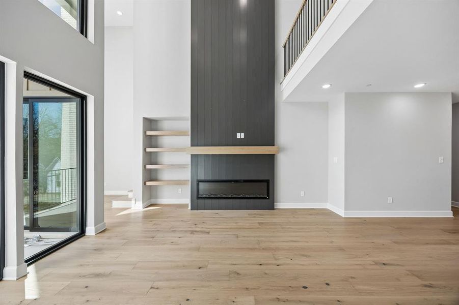 Unfurnished living room featuring a high ceiling, a large fireplace, light wood-type flooring, recessed lighting, and built in features Unfurnished living room featuring a high ceiling, a large fireplace, light wood-type flooring, recessed lighting, and built in features
