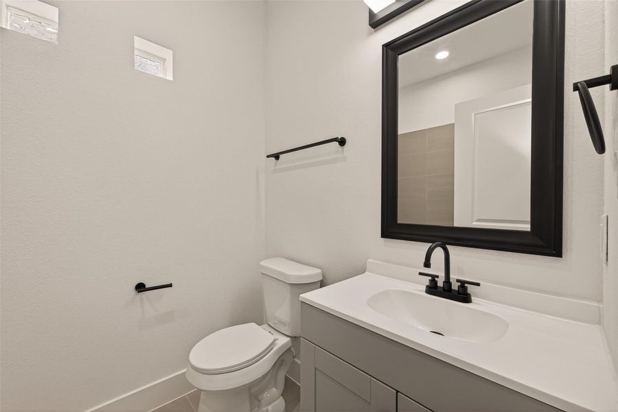 This bathroom features a clean, modern design with a white vanity and black fixtures. It includes a toilet and a large mirror above the sink, complemented by neutral tile flooring and two small windows for natural light. This bathroom features a clean, modern design with a white vanity and black fixtures. It includes a toilet and a large mirror above the sink, complemented by neutral tile flooring and two small windows for natural light.