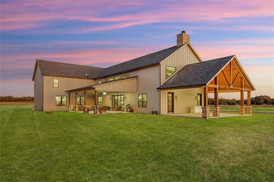 Back of house at dusk with a shingled roof, a patio area, a yard, and a chimney