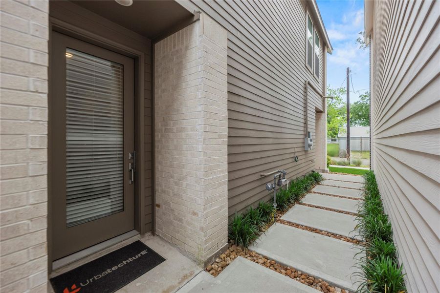 Stylish side entrance with a sleek glass-paneled door framed by neutral brick and siding. A charming walkway, lined with stepping stones and lush greenery, offers a warm and welcoming first impression.