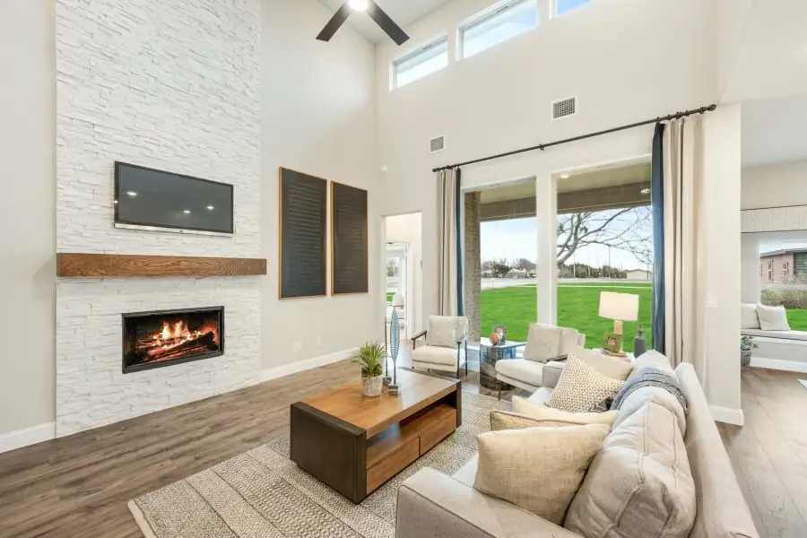 Living room with two-story ceiling, white stone fireplace, hardwood floors, and large windows overlooking a backyard.