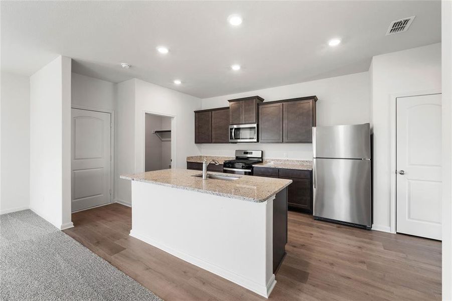 Kitchen featuring appliances with stainless steel finishes, dark brown cabinetry, light stone counters, a kitchen island with sink, and recessed lighting